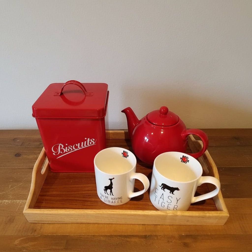 A red metal biscuit tin with white script lettering, a red teapot, and two white ceramic mugs with black dog silhouettes and text, all arranged on a wooden serving tray against a plain wall.