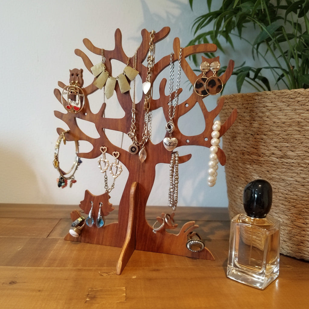 A wooden tree-shaped jewelry organizer displayed on a wooden table, holding various necklaces, earrings, and bracelets. A glass perfume bottle with a black cap sits to the right, with a woven basket and green plant in the background.