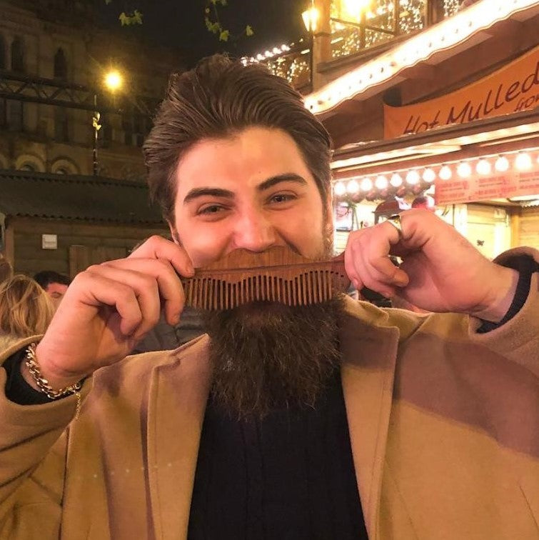 A man in a tan coat and black shirt holds a wooden comb over his beard, posing in front of a nighttime street market background with warm lights and a 'Hot Mulles' sign.