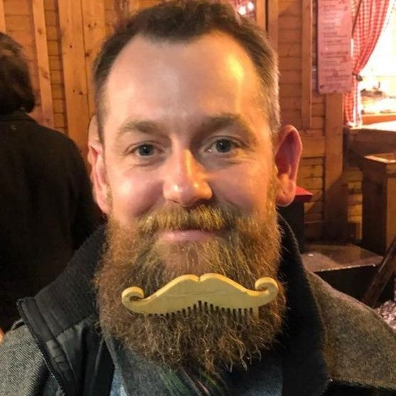 A man with a full brown beard wearing a small wooden mustache comb accessory in front of his mouth, photographed indoors in a wood-paneled room.