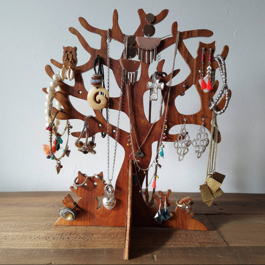 A wooden tree-shaped jewelry display stand made from a carved olive wood trunk, decorated with various necklaces, earrings, and rings, set on a wooden tabletop against a plain light gray wall.